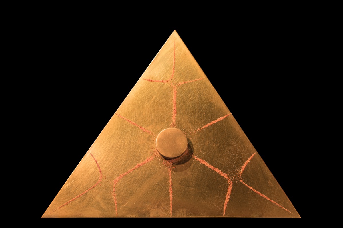 Placa triangular dorada con patrones simétricos dibujados por arena al vibrar con una frecuencia.